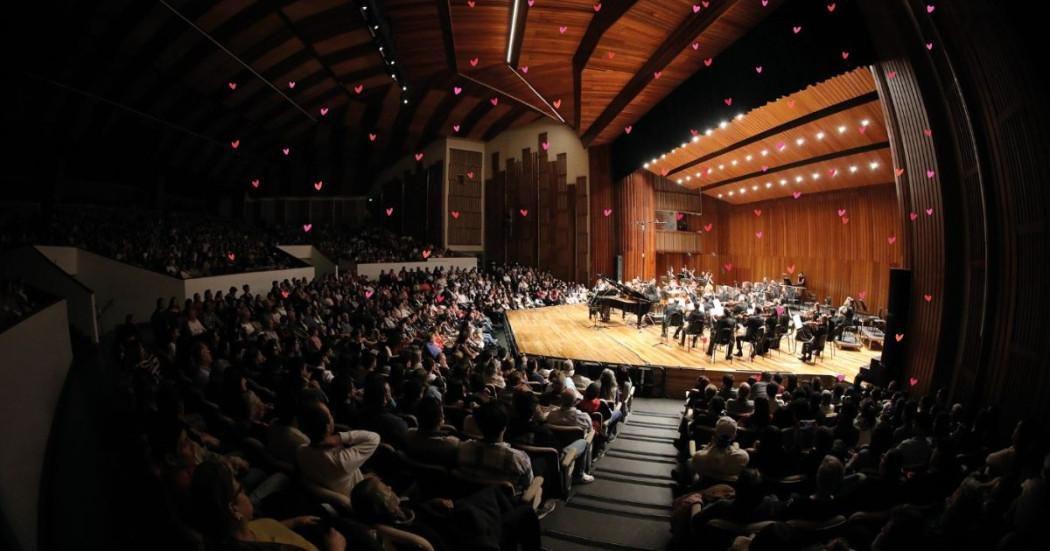 Auditorio León de Greiff de la Universidad Nacional de Colombia de arquitectura en madera y techo curvo, lleno de público sentado en graderías frente a un escenario iluminado. En el escenario se presenta una orquesta sinfónica junto a un piano de cola, con músicos distribuidos en semicírculo y un director al frente.
